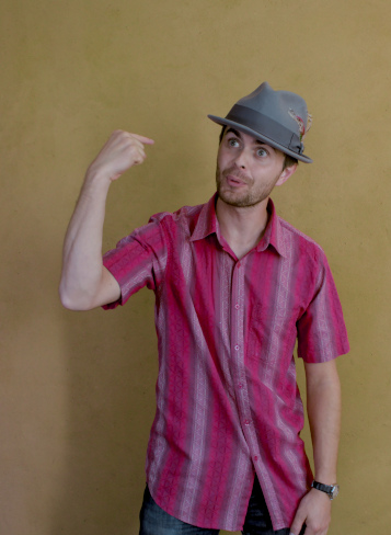 Un homme, chemise rouge, chapeau gris, regarde sa main comme si celle-ci lui parlait